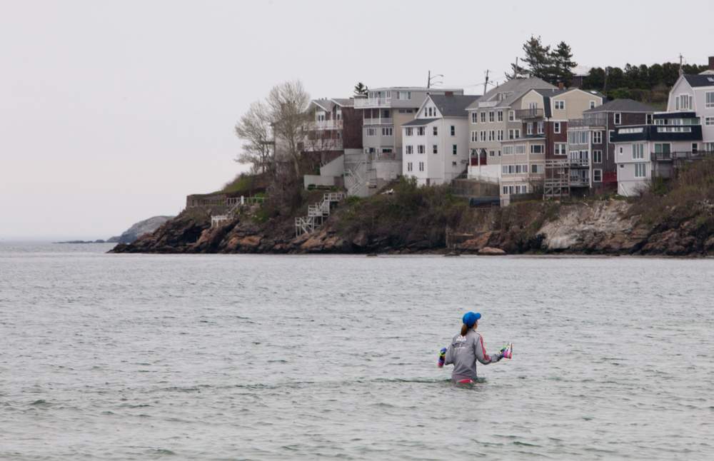 A person carries a pair of shoes while wading in the sea with Nahant homes visible in the distance.